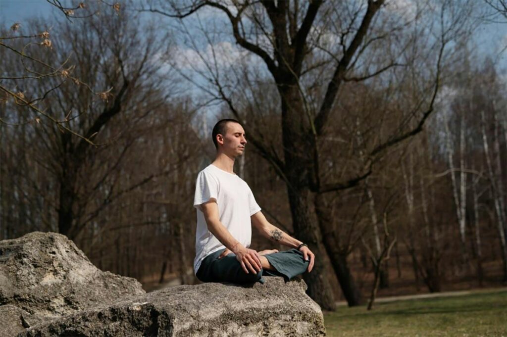 A person with a shaved head, wearing a white t-shirt and dark pants, is sitting cross-legged on a large rock outdoors. They appear to be meditating with their eyes closed. Bare trees and a partly cloudy sky are visible in the background, epitomizing a moment of peaceful reflection aligning with Progressive Mobility Physio & Performance principles.