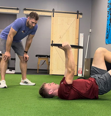 A man in a blue shirt and gray shorts observes another man in a red shirt and gray shorts who is lying on his back, lifting a weighted bar. They are in the Progressive Mobility Physio & Performance gym with green flooring and wooden doors in the background.
