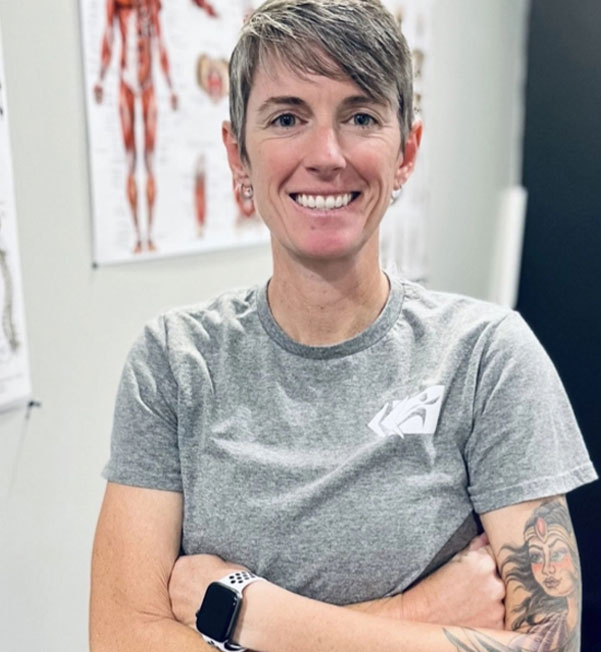 A person with short hair and a tattoo on their right arm stands smiling with arms crossed, wearing a grey t-shirt and a smartwatch. Anatomical muscle charts are seen in the background on a light-colored wall, hinting at the expertise of Progressive Mobility Physio & Performance.