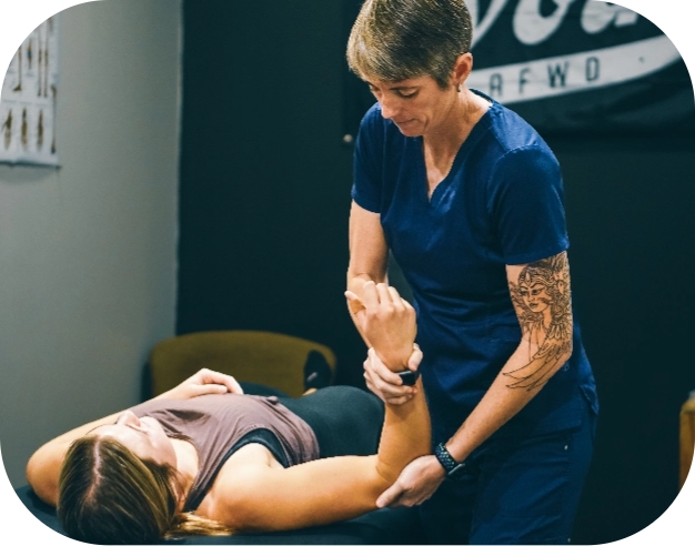 A therapist from Progressive Mobility, wearing blue scrubs, is assisting a woman lying on her back with exercises. The physio holds the woman's arm, guiding her through a physical therapy session in a professional setting.